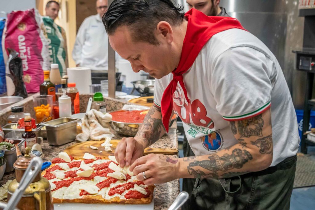 World Pizza Champion Tony Gemignani wears a ared scarf and places mozzarella on freshly baked grandma pizza